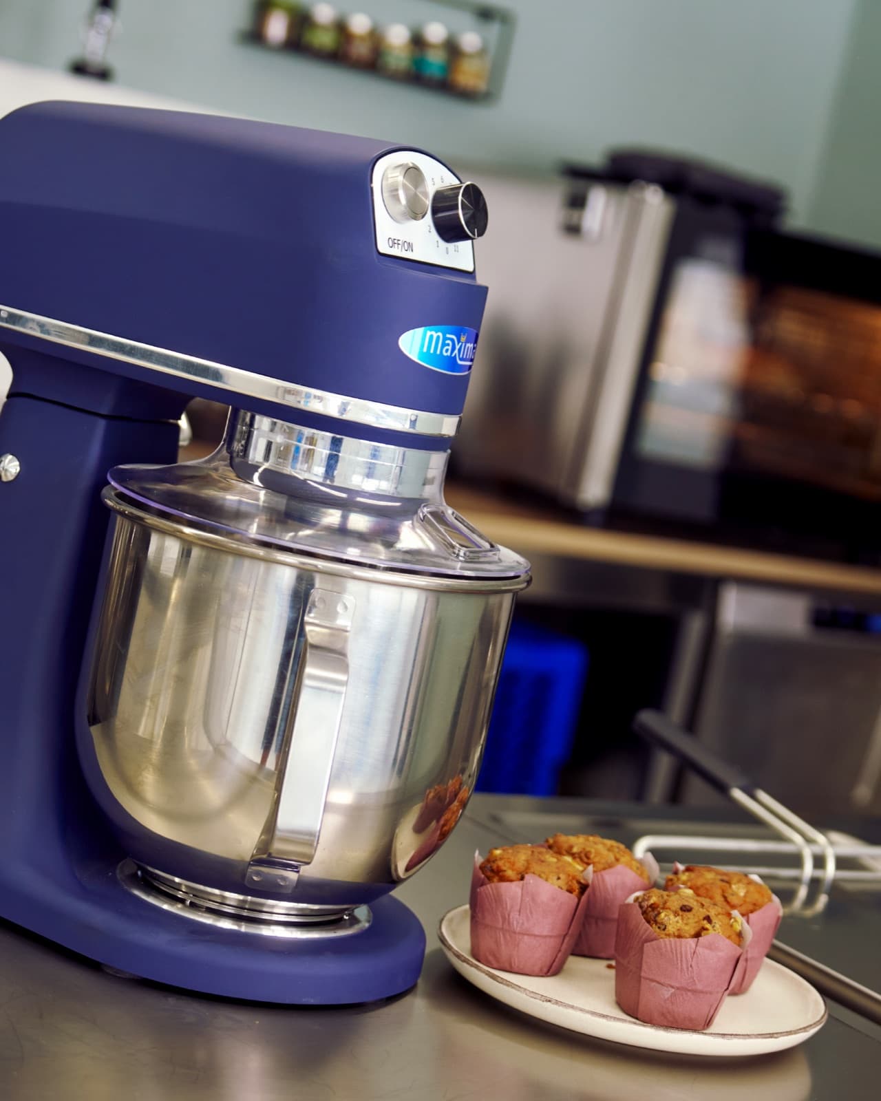 Een blauwe Maxima keukenmachine met roestvrijstalen kom in het atelier, naast een bordje met verse muffins.